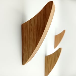 May include: Three wooden wall hooks with a natural oak finish are mounted on a white wall. The hooks have a curved, triangular design, providing a modern and minimalist aesthetic. The wood grain is visible.