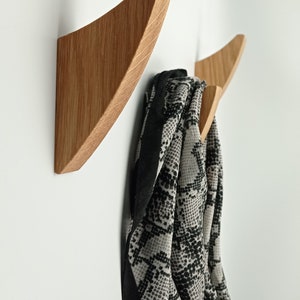 May include: Two wooden wall hooks with a natural finish. One hook is mounted on the wall, and the other is partially visible. A scarf with a black and white pattern hangs from the second hook. The hooks have a modern, minimalist design.
