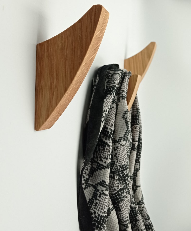 May include: Two wooden wall hooks with a natural finish. One hook is mounted on the wall, and the other is partially visible. A scarf with a black and white pattern hangs from the second hook. The hooks have a modern, minimalist design.
