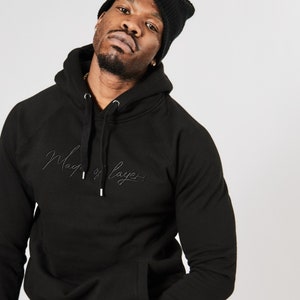 以下が含まれることがあります： 黒いフード付きスウェットシャツに「Mag of Layer」という文字が刺繍されています。モデルは黒いビーニーをかぶっています。