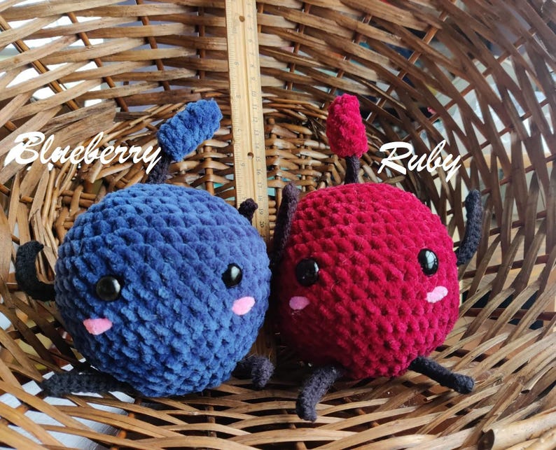 Peut inclure: Deux peluches en crochet en forme de myrtilles et de cerises. La myrtille est bleue avec des yeux noirs et des joues roses. La cerise est rouge avec des yeux noirs et des joues roses. Les jouets sont dans un panier en osier. La myrtille est &eacute;tiquet&eacute;e "Blueberry" et la cerise est &eacute;tiquet&eacute;e "Ruby".