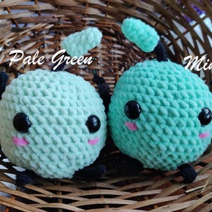 Peut inclure: Deux jouets en peluche crochet&eacute;s en forme de cr&eacute;ature ronde avec de grands yeux et une petite bouche. Le jouet de gauche est vert p&acirc;le et le jouet de droite est vert menthe. Les jouets sont assis dans un panier en osier.