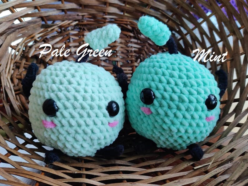 Peut inclure: Deux jouets en peluche crochet&eacute;s en forme de cr&eacute;ature ronde avec de grands yeux et une petite bouche. Le jouet de gauche est vert p&acirc;le et le jouet de droite est vert menthe. Les jouets sont assis dans un panier en osier.