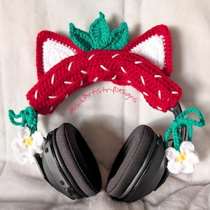May include: Black headphones decorated with a crocheted strawberry design. The red strawberry top has white accents and green leaves. White flowers with yellow centres and green vines are attached. The text "@EsolArtistryDesigns" is visible.
