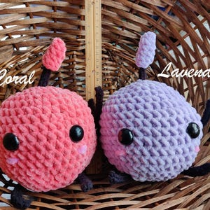 Peut inclure: Deux jouets en peluche crochet&eacute;s en forme de cerise et de fleur de lavande. La cerise est rose corail avec des yeux noirs et une petite bouche rose. La fleur de lavande est violette avec des yeux noirs et une petite bouche rose. Les deux jouets sont assis dans un panier en osier.