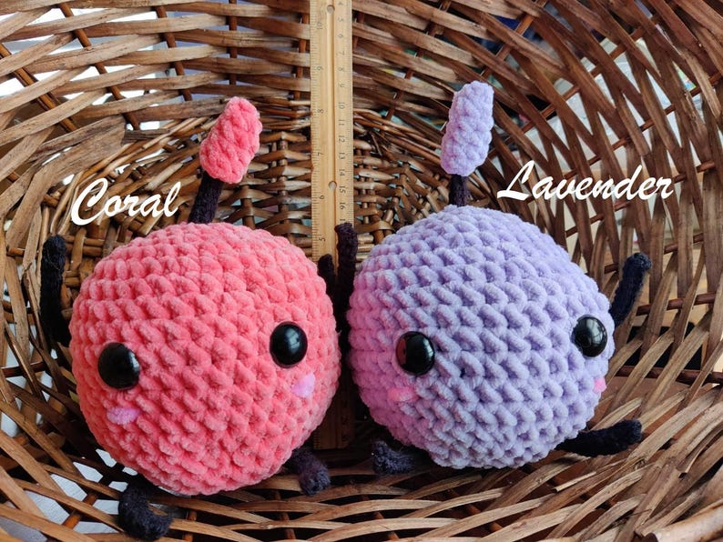 Peut inclure: Deux jouets en peluche crochet&eacute;s en forme de cerise et de fleur de lavande. La cerise est rose corail avec des yeux noirs et une petite bouche rose. La fleur de lavande est violette avec des yeux noirs et une petite bouche rose. Les deux jouets sont assis dans un panier en osier.