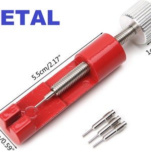 May include: Red metal watch band tool with a silver knob and three silver pins. The tool measures 5.5 inches long and 1.5 inches wide. The knob measures 1 inch in diameter. METAL is written in blue letters at the top of the image.