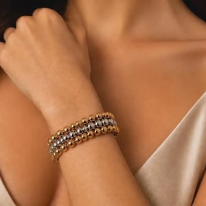 May include: A multi-strand beaded bracelet featuring three rows of polished gold-tone spherical beads and one central row of polished silver-tone spherical beads, all arranged in a stacked design on a flexible cord.