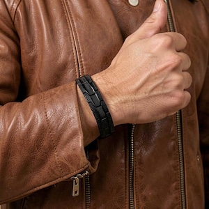 May include: A matte black link bracelet featuring a textured, geometric pattern on each rectangular segment. The bracelet is worn on the wrist, showcasing a dark, industrial aesthetic with a repeating grid-like design across the entire band.
