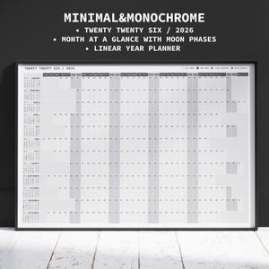 Puede incluir: Un planificador anual minimalista y monocromático de 2026 enmarcado. El calendario muestra una vista mensual con fases lunares. El texto superior dice "MINIMAL&MONOCHROME" y "TWENTY TWENTY SIX / 2026". El planificador está sobre una superficie de madera blanca.
