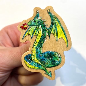 May include: A wooden dragon-shaped pin with a green body, yellow belly, and blue accents. The dragon has yellow and green wings and is exhaling a red flame. The pin is made of light-coloured wood.