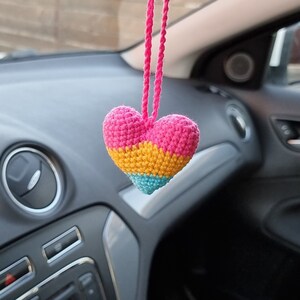May include: A crocheted heart-shaped car charm with pink, yellow and blue stripes hanging from a rear view mirror in a car.