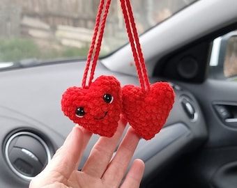 May include: Two red crocheted hearts with smiling faces hanging from a rearview mirror in a car.
