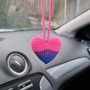 May include: A crocheted heart-shaped car charm in pink, purple and blue colours. The charm is hanging from the rear-view mirror of a car.