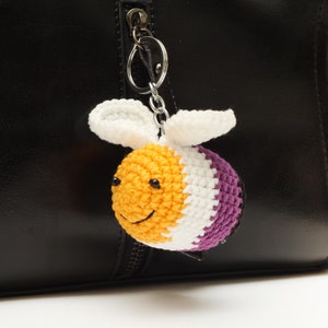 以下が含まれることがあります： 黄色、白、紫のストライプのクロシェ編みビーキーチェーン。ビーは笑顔で、シルバーのキーリングに付けられています。