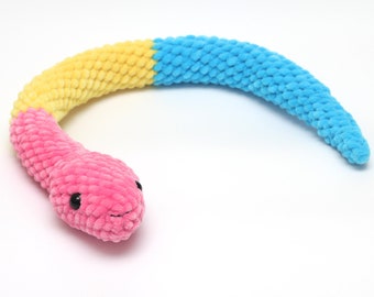 Puede incluir: Un juguete de peluche con forma de serpiente en rosa, amarillo y azul. La cabeza de la serpiente es rosa, pasando a amarillo y luego a azul. El juguete tiene pequeños ojos negros y está hecho de un material suave y texturizado.
