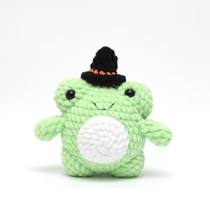 May include: A green crocheted frog wearing a black witch hat with orange trim. The frog has white yarn for its belly and black eyes.
