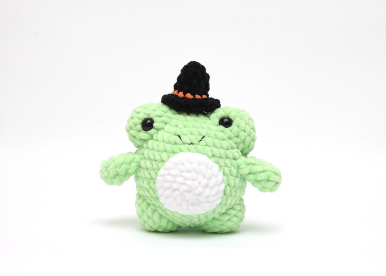 May include: A green crocheted frog wearing a black witch hat with orange trim. The frog has white yarn for its belly and black eyes.