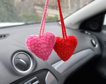 May include: Two crocheted heart-shaped ornaments hanging from a rearview mirror in a car. The heart on the left is pink and the heart on the right is red.