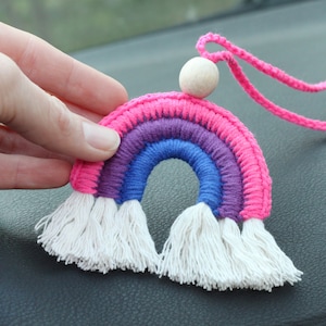 以下が含まれることがあります： 虹色の毛糸で編まれた車のチャーム。白い房飾り、木製のビーズ付き。ピンクの毛糸で編まれた紐で吊り下げられています。