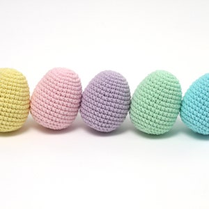 Könnte beinhalten: Fünf handgefertigte Häkeleier in Pastellfarben: Gelb, Rosa, Lila, Grün und Blau. Jedes Ei hat eine strukturierte Oberfläche, die durch die Häkelstiche erzeugt wird. Die Eier sind in einer Reihe vor weißem Hintergrund angeordnet.