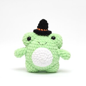 May include: A handmade, light green crochet frog plushie with a white belly and black eyes. The frog is wearing a small black witch hat with orange trim, perfect for Halloween or a whimsical gift.