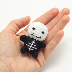 May include: A small crocheted skeleton toy with a white head and black body. The skeleton has a smiling face with black eyes and a black mouth.