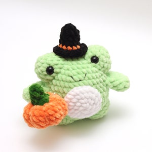 May include: A green crocheted frog wearing a black witch hat and holding an orange crocheted pumpkin.