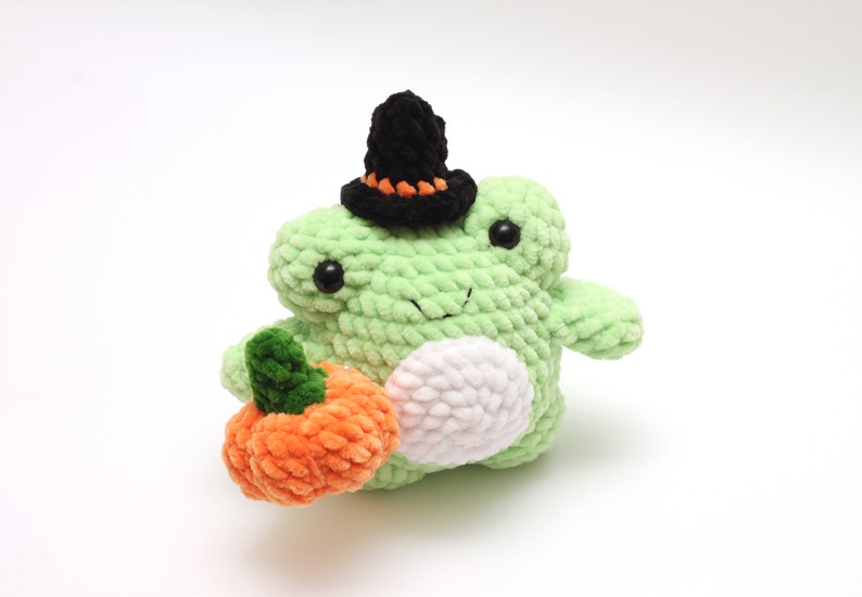 May include: A green crocheted frog wearing a black witch hat and holding an orange crocheted pumpkin.