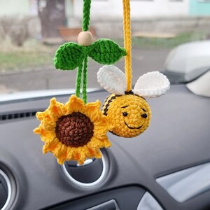 May include: A crocheted sunflower and bee car charm hanging from a rearview mirror. The sunflower is yellow with a brown centre and the bee is yellow with black stripes and white wings.