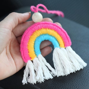 Può includere: Un ciondolo per auto a forma di arcobaleno colorato con nappe bianche e una perla di legno. L'arcobaleno è fatto di filo rosa, giallo, blu e bianco.