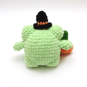 May include: A green crocheted plush toy wearing a black witch hat with an orange brim. The toy is sitting on a white background.