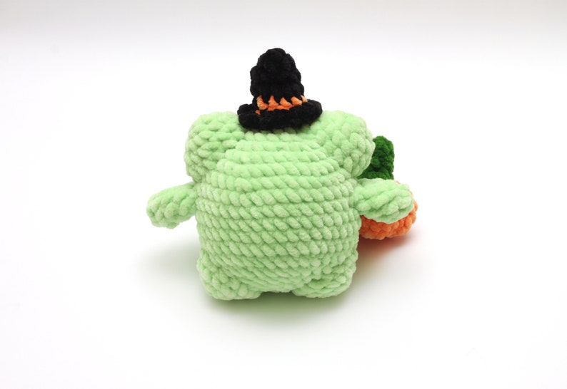 May include: A green crocheted plush toy wearing a black witch hat with an orange brim. The toy is sitting on a white background.