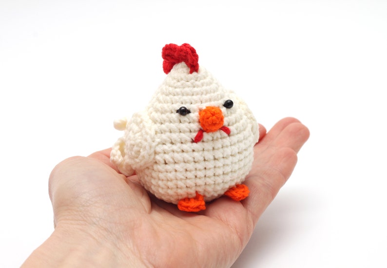 K&ouml;nnte beinhalten: Ein wei&szlig;es H&auml;keltier in Form eines Huhns mit rotem Kamm, orangem Schnabel und schwarzen Augen. Das Huhn sitzt in der Handfl&auml;che.