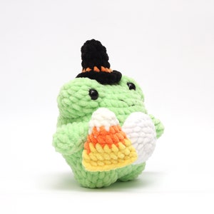 May include: A green crocheted frog wearing a black and orange witch hat, holding a candy corn and a white egg-shaped item. The frog has black eyes and is made of a soft, textured yarn. A Halloween-themed plush toy.
