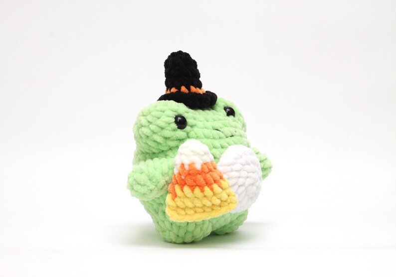 May include: A green crocheted frog wearing a black and orange witch hat, holding a candy corn and a white egg-shaped item. The frog has black eyes and is made of a soft, textured yarn. A Halloween-themed plush toy.