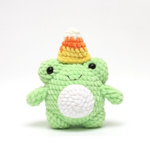 May include: A green crocheted frog wearing a yellow, orange, and white candy corn hat. The frog has a white belly and black eyes.