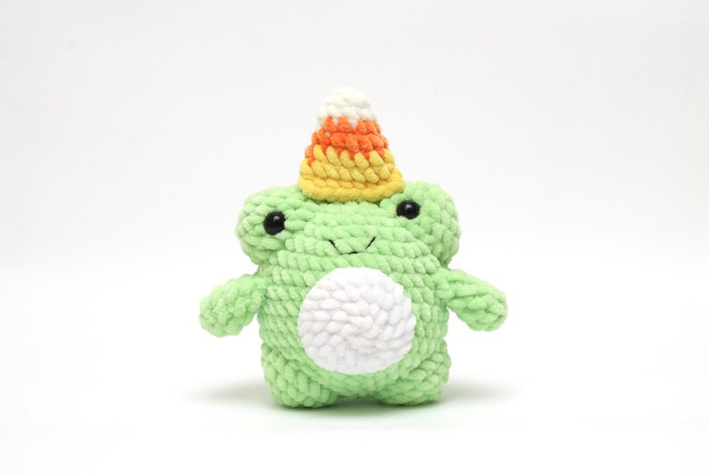 May include: A green crocheted frog wearing a yellow, orange, and white candy corn hat. The frog has a white belly and black eyes.
