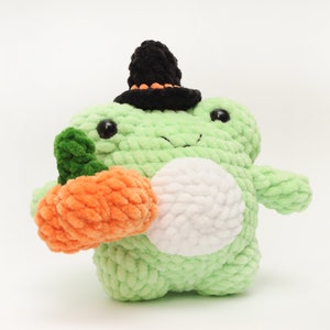 May include: A green crocheted frog wearing a black witch hat with an orange trim, holding a crocheted orange pumpkin.