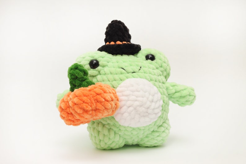 May include: A green crocheted frog wearing a black witch hat with an orange trim, holding a crocheted orange pumpkin.