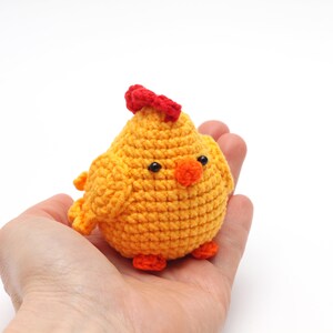 K&ouml;nnte beinhalten: Ein kleines geh&auml;keltes H&uuml;hner-Spielzeug mit einem roten Kamm, einem orangefarbenen Schnabel und schwarzen Augen. Das K&uuml;ken ist gelb und sitzt in der Hand einer Person.