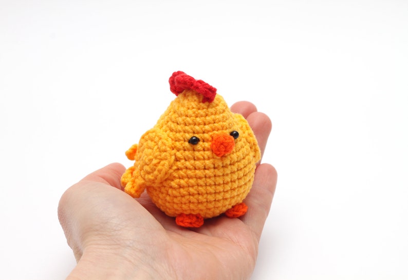 K&ouml;nnte beinhalten: Ein kleines geh&auml;keltes H&uuml;hner-Spielzeug mit einem roten Kamm, einem orangefarbenen Schnabel und schwarzen Augen. Das K&uuml;ken ist gelb und sitzt in der Hand einer Person.
