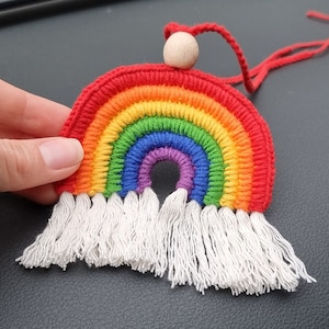 May include: A rainbow-coloured crocheted wall hanging with white fringe and a wooden bead at the top. The hanging is attached to a red string.