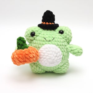 May include: A handmade, light green crochet frog wearing a black witch hat with an orange band. The frog holds an orange pumpkin with a green stem. The frog has a white belly and black eyes. A Halloween-themed plushie.