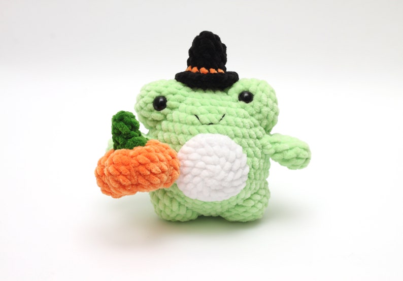 May include: A handmade, light green crochet frog wearing a black witch hat with an orange band. The frog holds an orange pumpkin with a green stem. The frog has a white belly and black eyes. A Halloween-themed plushie.