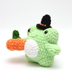 May include: A green crocheted frog wearing a black witch hat with orange accents holds a crocheted orange pumpkin.