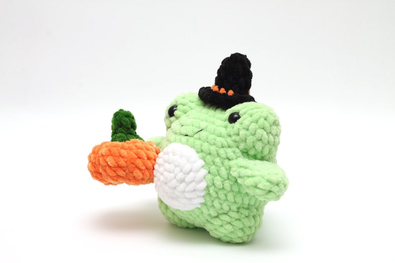 May include: A green crocheted frog wearing a black witch hat with orange accents holds a crocheted orange pumpkin.