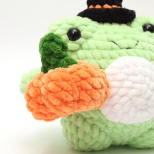 May include: A green crocheted frog wearing a black witch hat holds an orange crocheted pumpkin.