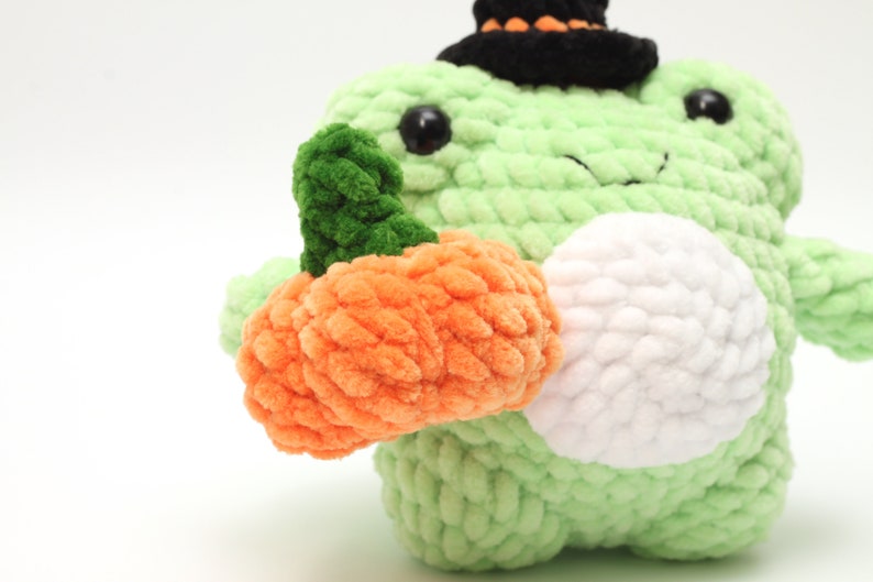 May include: A green crocheted frog wearing a black witch hat holds an orange crocheted pumpkin.