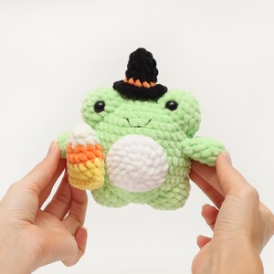 May include: A handmade, light green crochet frog wearing a black witch hat with orange trim. The frog holds a candy corn in its hand. The frog has a white belly and black eyes. A Halloween-themed plush toy.